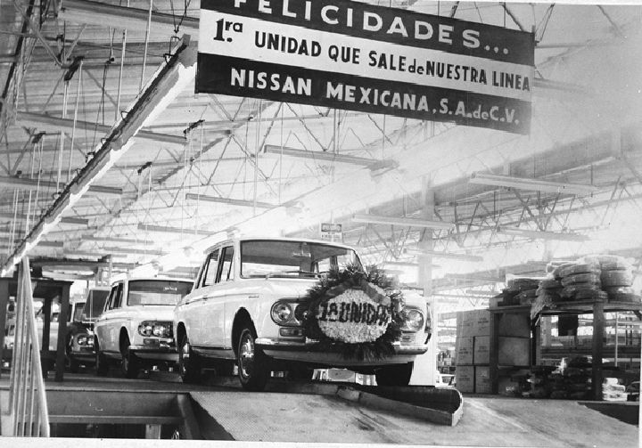Foto del recuerdo de la primera unidad que salió de la línea de producción de planta Civac, es un Datsun blanco. De fondo se ve la Planta.