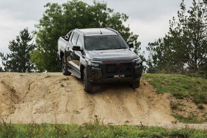 2026 Nissan Navara PRO-4X finished in Boulder Grey shown in a diagonal descent scene leaning down a sandy face bordered by tufts of grass, bushes and dark tyres.