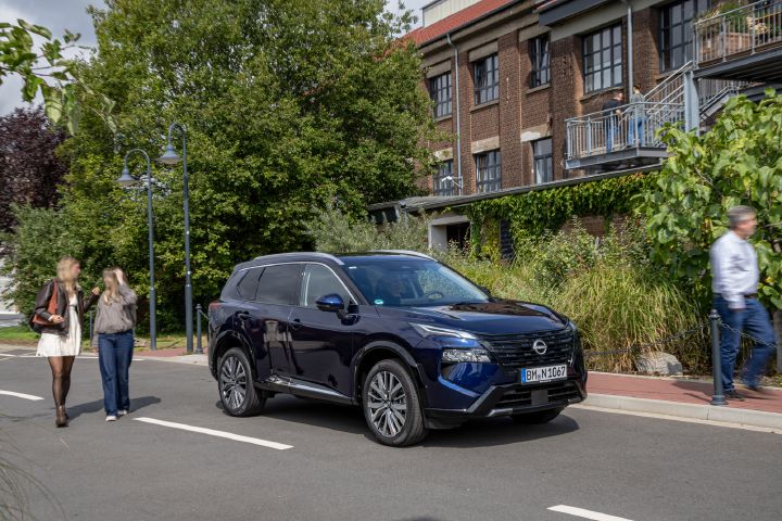 Blick von schräg vorne auf einen dunkelblauen Nissan X‑Trail, der am Straßenrand steht, um das Fahrzeug herum laufen drei Personen