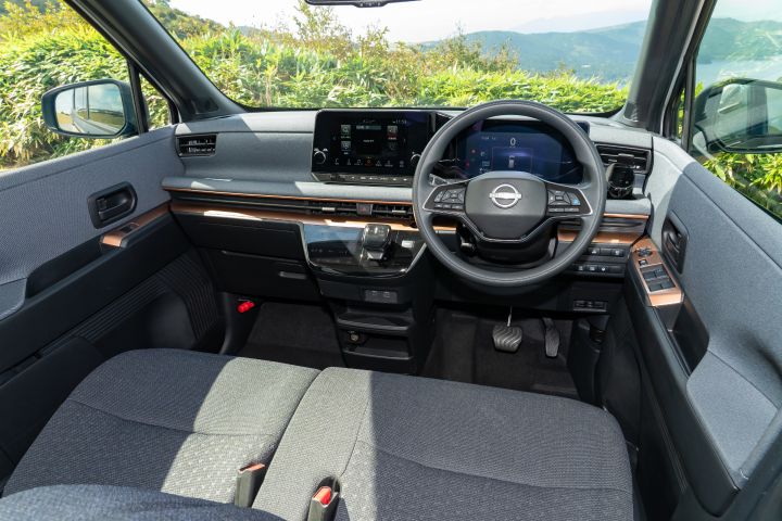 Interior view of a Nissan SAKURA from the front, showing a black steering wheel and digital instrument cluster in the center. The dashboard features a large display and control panel, with green shrubs and mountains visible outside.