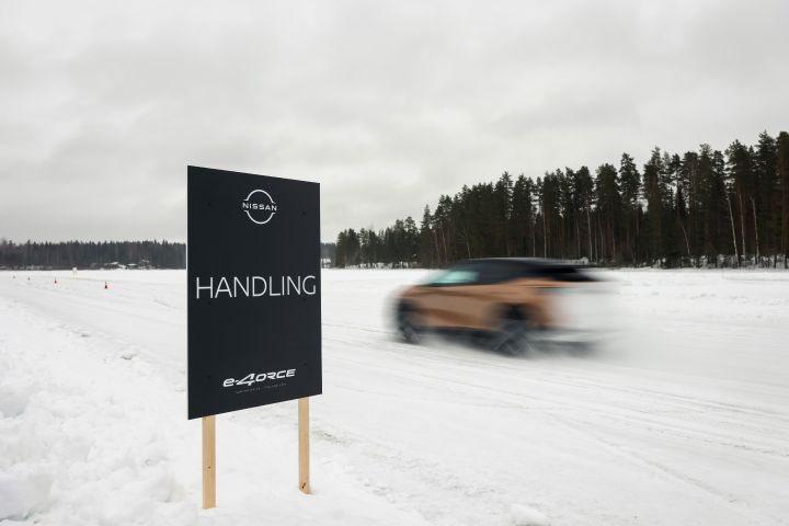 Schwarzes Schild mit der Aufschrift Nissan e‑4ORCE Handling im Vordergrund, dahinter fährt unscharf das Elektroauto Nissan Ariya e‑4ORCE in Akatsuki Copper mit Dach und Außenspiegel in Pearl Black auf Schnee vorbei