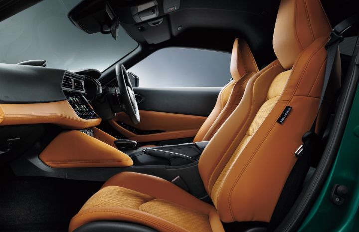 Interior view of a Fairlady Z featuring a tan cabin, photographed from the rear passenger-side area looking forward. Tan sport seats dominate the foreground, showcasing their pronounced side bolsters and sculpted design. Matching tan leather trim extends across the dashboard and door panels, contrasting with the black center console and instrument panel. The right-hand-drive steering wheel and center infotainment display are visible, emphasizing the sporty yet premium cockpit layout. A portion of the exterior Uryu Green body color can be seen along the door opening.