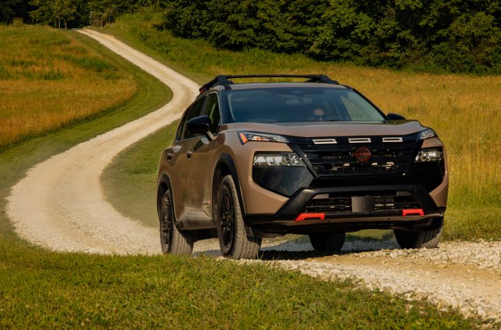 Front right 3/4 angle of a 2025 Nissan Rogue Rock creek driving up a small dirt road through a field with trees behind.