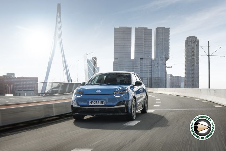 En blå Nissan MICRA, set ¾ forfra kører over en bro med en by I baggrunden og et Årets Bil i Danmark logo nederst til højre