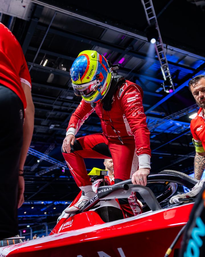 Oliver Rowland, der einen roten Rennanzug und einen bunten Helm trägt, steigt in seinen rot-weißen Nissan Formel-E-Rennwagen ein. Teammitglieder stehen in der Nähe, im Hintergrund sind eine Halle und helle Deckenbeleuchtung zu sehen.