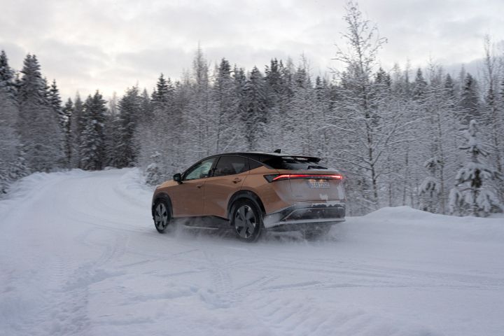 Elektroauto Nissan Ariya e‑4ORCE in Akatsuki Copper mit Dach und Außenspiegel in Pearl Black fährt auf schneebedeckter Straße mit verschneiten Bäumen auf beiden Seiten, 3/4 Heck