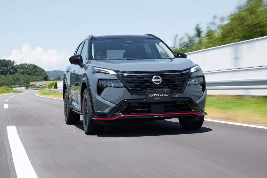 A grey Nissan X-Trail NISMO is driving on a road. The vehicle is captured from the front and appears to be in motion. The background features trees and a guardrail, and the license plate reads “NISSAN.”