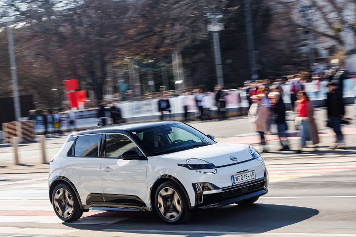 Bei sonnigem Wetter fährt ein weißer Nissan Micra mit schwarzem Kontrastdach über eine Straße, rundherum sind verschwommen viele Menschen zu sehen