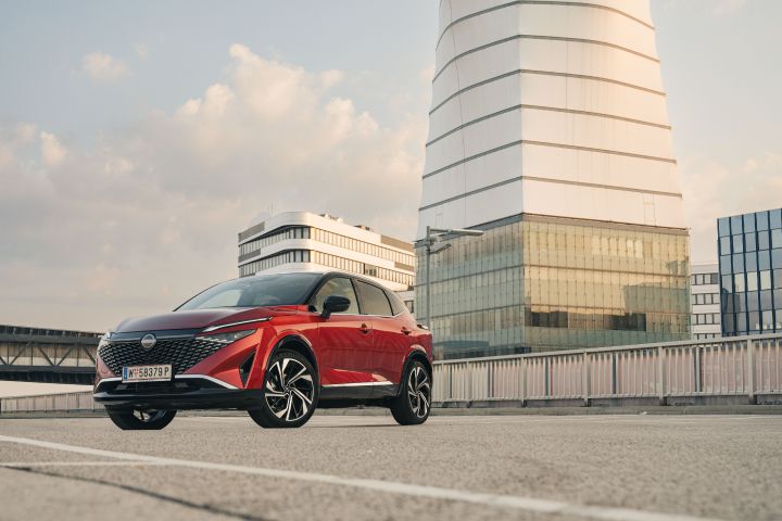 Ein Nissan Qashqai in Fuji Sunset Red steht schräg auf einem Parkplatz vor einem Hochhaus