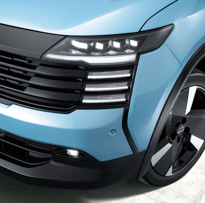 Close-up of the front of a bluish Nissan Kicks. The vehicle faces right, highlighting the horizontal design of the grille and headlights.