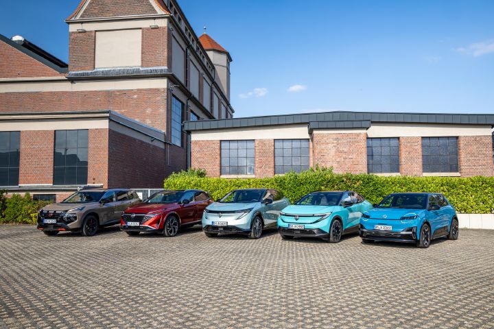 Vor einem Backsteingebäude und einer grünen Hecke stehen nebeneinander die fünf Nissan Modelle Nissan X‑Trail N-Trek, Nissan Qashqai, Nissan Ariya, Nissan Leaf und Nissan Micra, Blick von schräg vorne aus größerer Entfernung