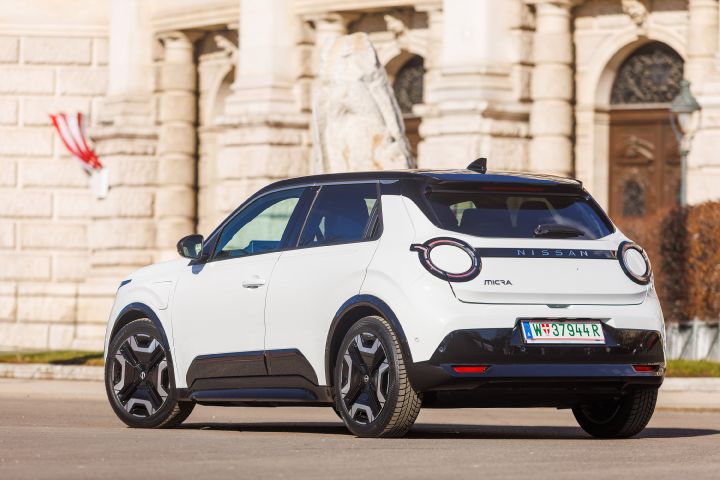 Blick von schräg hinten auf einen weißen Nissan Micra mit schwarzem Kontrastdach, der in der Sonne vor einem historischen Gebäude mit Österreich-Flagge steht