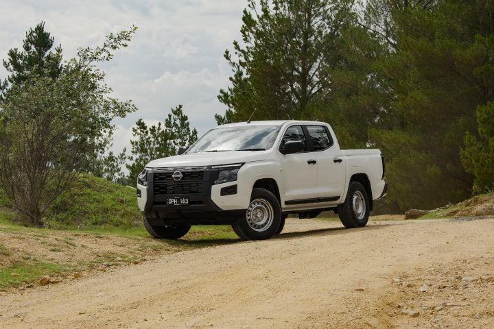 2026 Nissan Navara SL dual cab ute finished in Alpine White presented from the front-left on a bush-lined gravel road, with the steel wheels and upright body clearly visible.