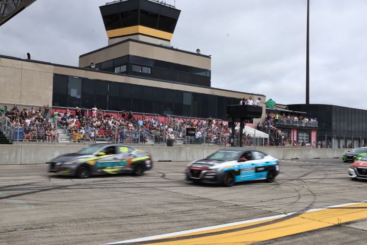 Sentra race cars speeding past a grandstand filled with spectators, under an overcast sky.