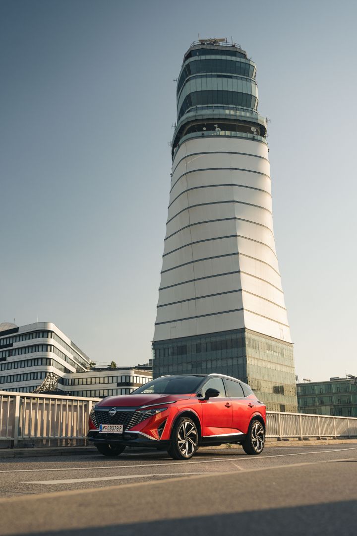 Vor einem hohen weißen Turm steht ein Nissan Qashqai in Fuji Sunset Red auf einem Parkplatz