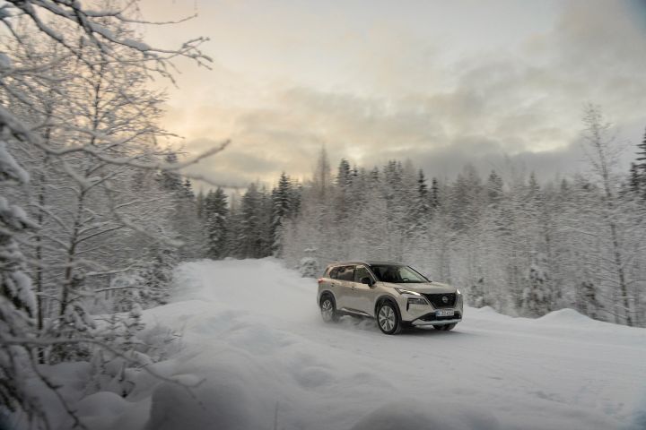 Silberner Nissan X‑Trail e‑Power e‑4ORcE fährt auf schneebedeckter Straße mit verschneiten Bäumen auf beiden Fahrbahnseiten, 3/4 Front