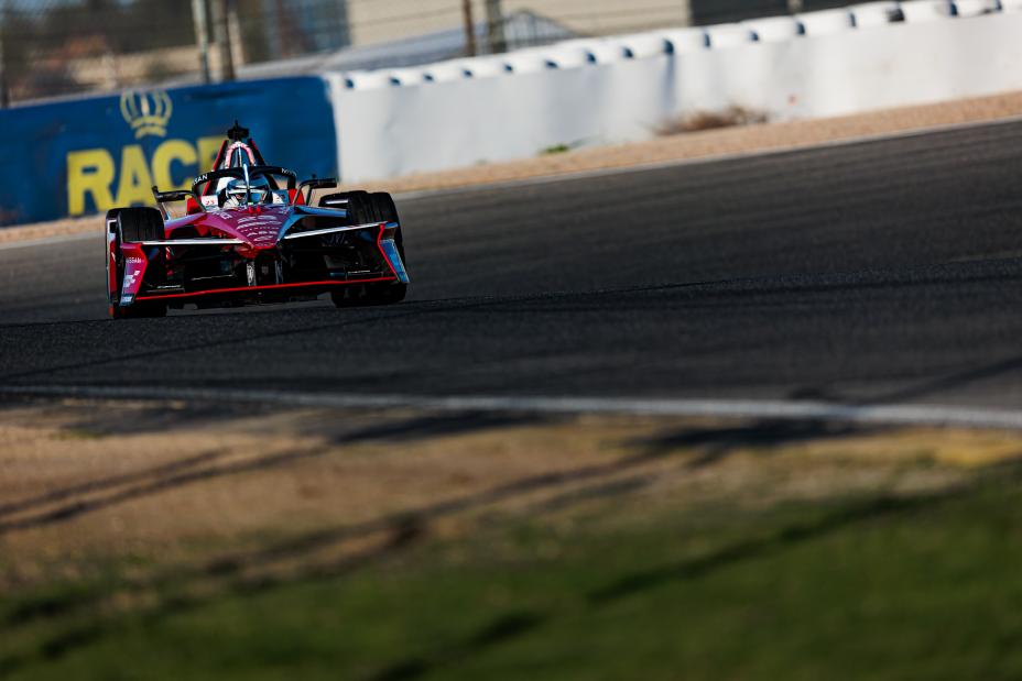 Oliver Rowland shown in the top-left of the shot with a blue sponsorship barrier just behind him as he exits the corner with sand and grass also shown at the bottom of the photo.