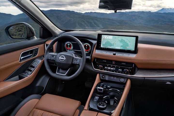 Der Innenraum des neuen 2026 Nissan X‑Trail Tekna konzentriert sich auf den Fahrersitz, das Lenkrad und das Armaturenbrett mit einem digitalen Kombiinstrument und einem großen Touchscreen, auf dem eine Navigationskarte angezeigt wird. Der Innenraum ist mit kastanienbrauner Premium-Stepplederpolsterung, edlen braunen Holzverkleidungen und verschiedenen Bedientasten am Lenkrad und an der Mittelkonsole ausgestattet, die ein modernes und luxuriöses Design unterstreichen.