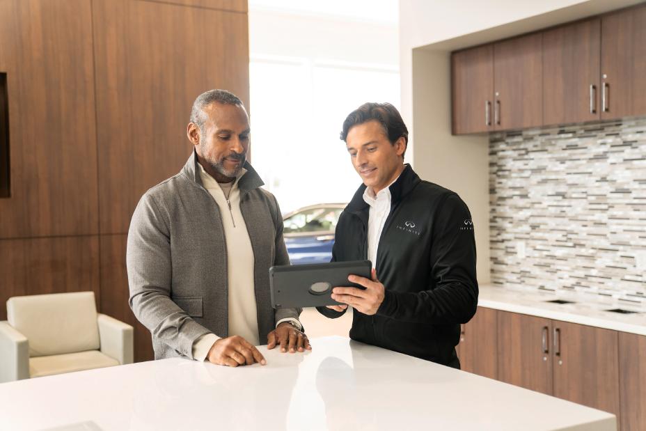 An INFINITI retail member in black jacket shows a client in grey jacket information on a tablet.
