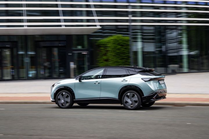 Blick von schräg hinten auf einen fahrenden hellblauen Nissan Ariya vor einem Bürogebäude