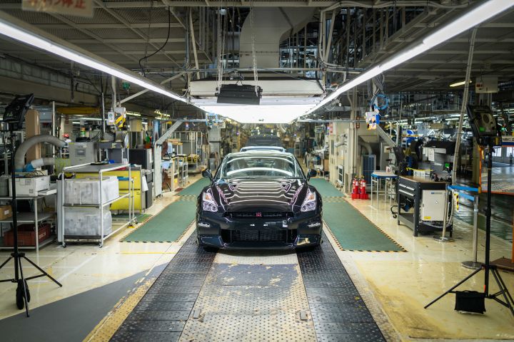 日産自動車 栃木工場の製造ラインにて、ミッドナイトパープルのR35 GT-Rが前景に配置されており、後方には青色の車両を含む複数の車が並んでいる。工場内は明るい照明に照らされ、周囲には自動車製造に必要な機械設備が整っており、高性能車の組立工程が描かれている。
