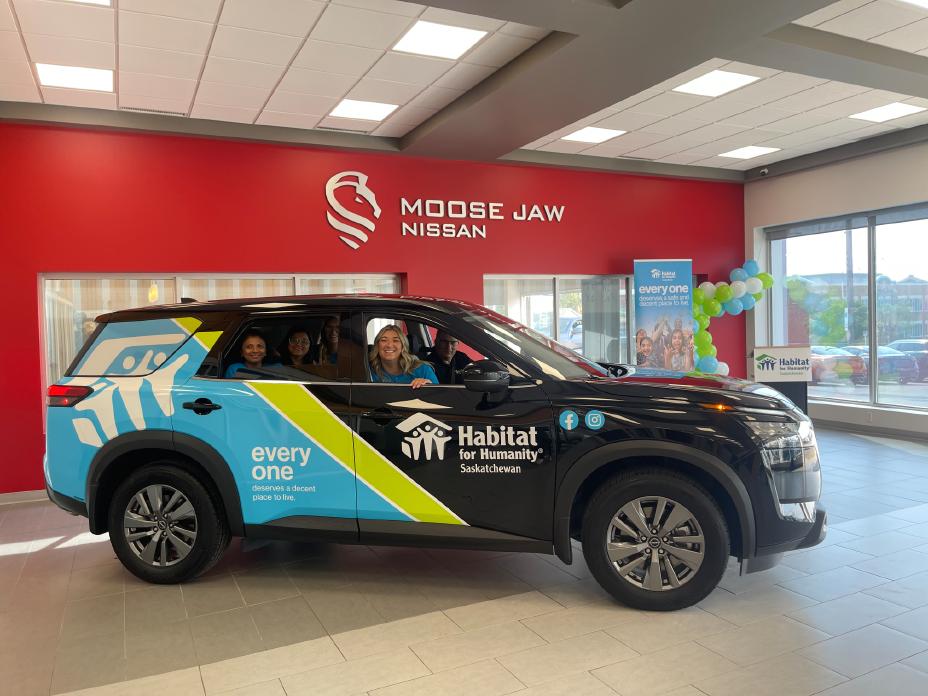 A 2024 Nissan Pathfinder wrapped in Habitat for Humanity Saskatchewan branding is parked inside a dealership. The vehicle has a blue, black, and green design with the Habitat for Humanity logo and the slogan "every one deserves a decent place to live." Five people are smiling inside, with a red wall in the background displaying "Moose Jaw Nissan."