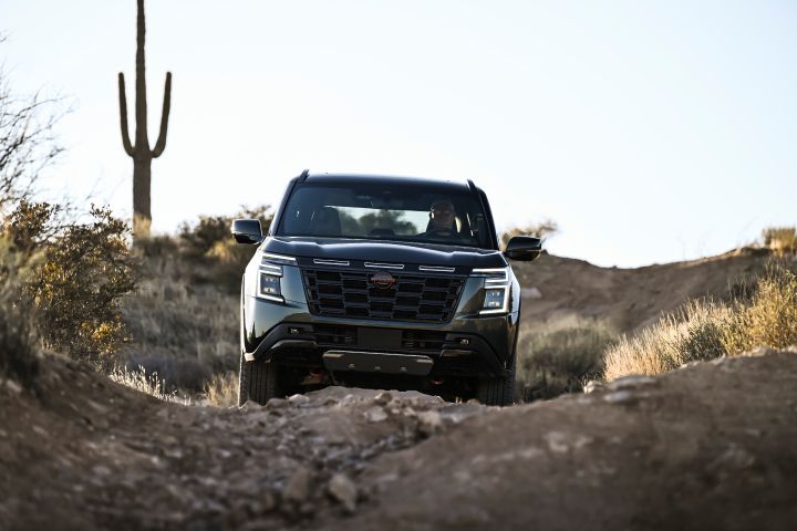 Front view of a 2025 Nissan Armada PRO-4X driving on uneven, rocky terrain during sunset, a cactus is behind.