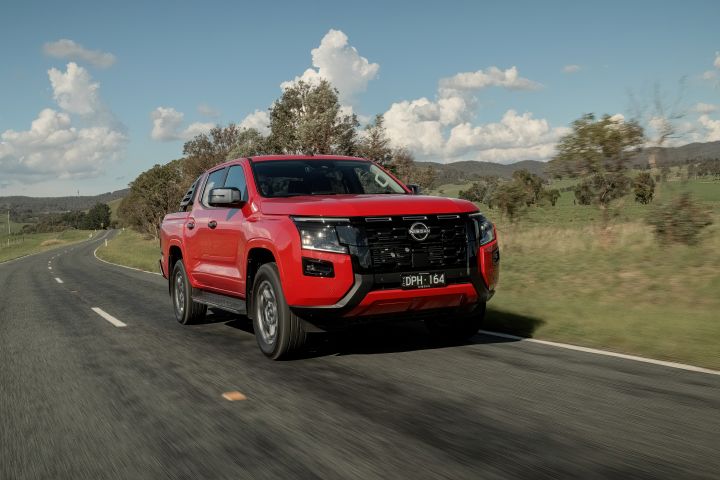 Aerial view of a 2026 Nissan Navara ST‑X dual cab finished in Outback Red, travelling on a winding rural road bordered by dry grass and scattered shrubs.