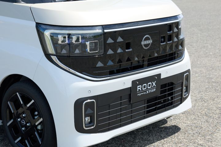 The front of the new Nissan Roox kei car, in white with a distinctive black grille and headlights, is photographed from the left side toward the front-right angle. The background features an asphalt surface.