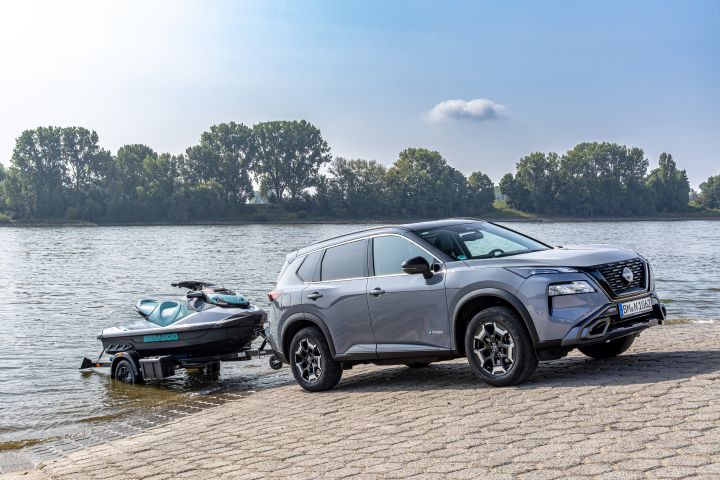 Ein metallicgrau lackierter Nissan X‑Trail N-Trek mit Bootsanhänger steht bei Sonnenschein schräg an einer gepflasterten Rampe am Fluss Rhein, der Bootsanhänger mit Jetski steht im Wasser