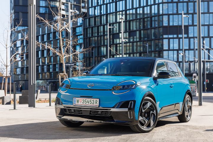 Vor einem hohen Bürogebäude mit gläserner Fassade steht ein leuchtend blauer Nissan Micra mit schwarzem Kontrastdach, 7/8-Frontansicht