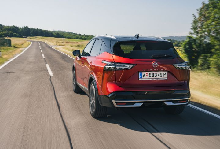 Fahraufnahme von schräg hinten eines Nissan Qashqai in Fuji Sunset Red auf einer sonnigen, geraden Landstraße