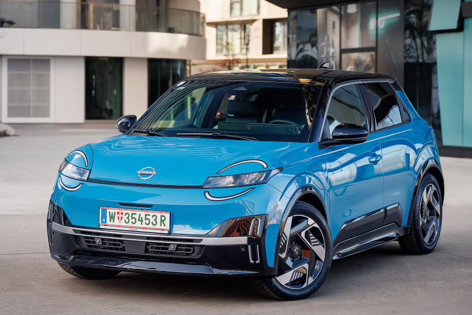 3/4-Frontansicht eines leuchtend blauen Nissan Micra mit schwarzem Kontrastdach, der vor zwei modernen Gebäuden steht