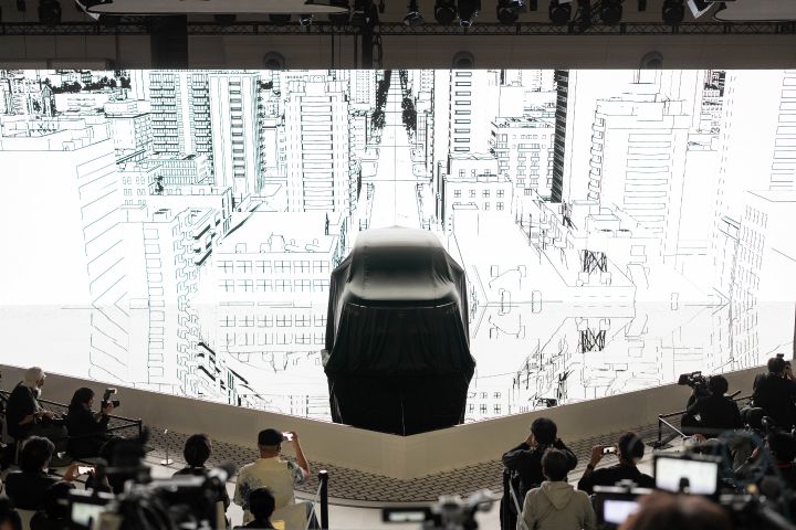 At the center of the stage, the all-new Elgrand is covered with a black cloth. The background features a white screen with a cityscape line drawing. In the foreground, members of the press are gathered, taking photos with cameras.