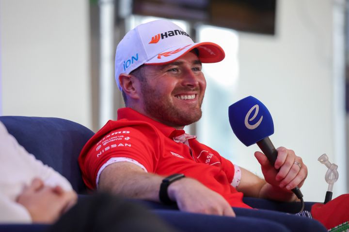 Oliver Rowland lächelt mit einer weißen Hankook-Kappe auf dem Kopf, während er ein rotes Polohemd des Nissan Formula E Teams trägt und ein Mikrofon in der linken Hand hält.
