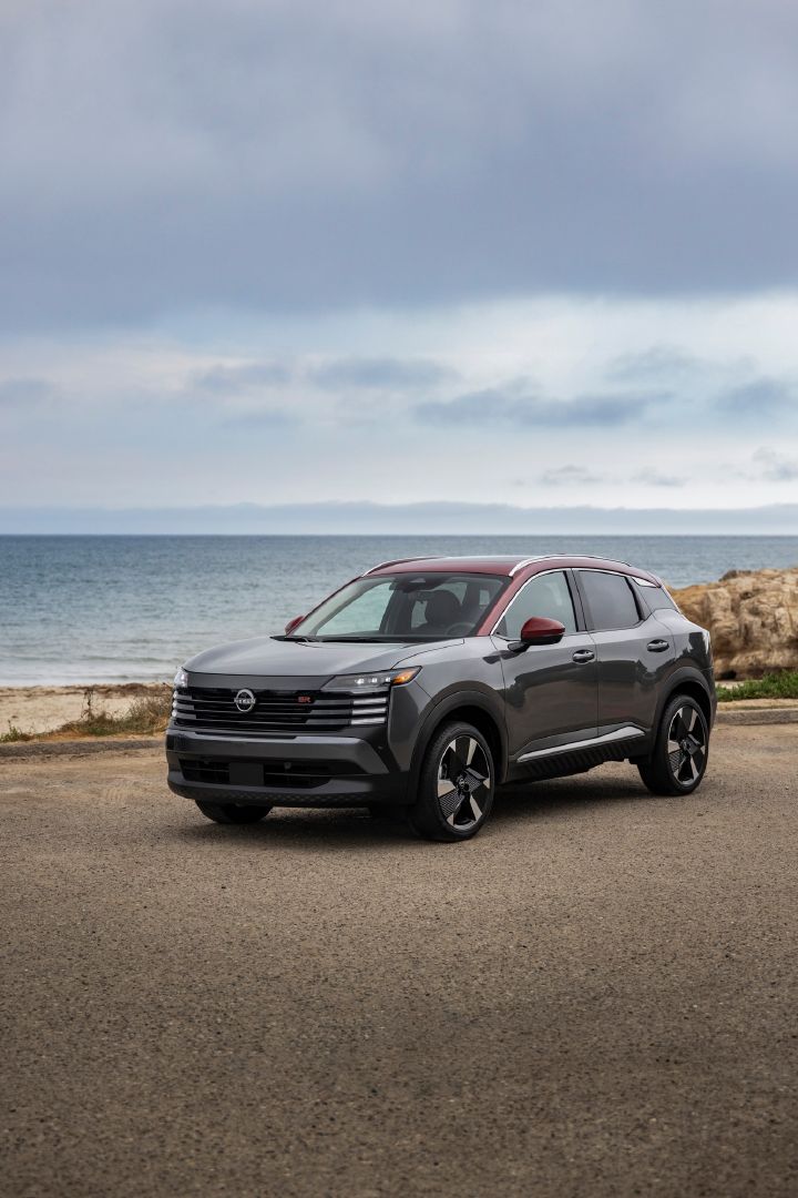 Front left 3/4 view of a silver 2025 Nissan Kicks parked on a gravel lot overlooking the ocean on a cloudy day.