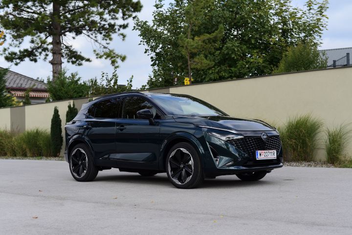 3/4 Frontansicht eines dunkelgrünen Nissan Qashqai e‑Power, der vor einer hellen Mauer am Straßenrand steht, dahinter ragen Bäume auf