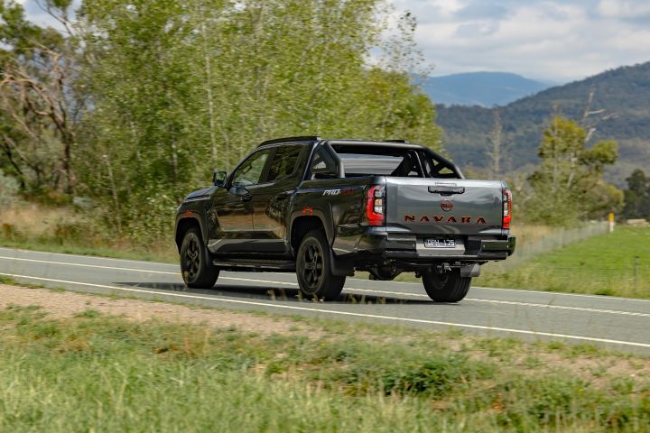 2026 Nissan Navara PRO-4X finished in Boulder Grey viewed from the rear three-quarter angle while departing along a two-lane road with sports bar and tow hitch.