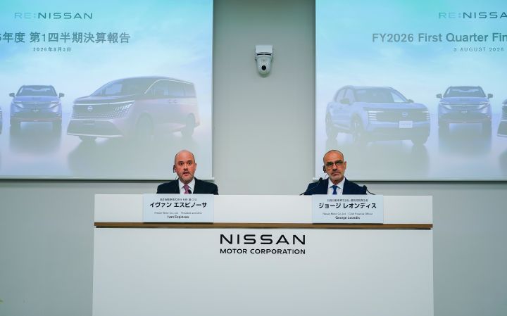 In a conference setting with a white backdrop, two people are seated side by side behind a podium. Large screens in the background display vehicle images and presentation slides, shown from a front-facing view during a press conference or presentation.
