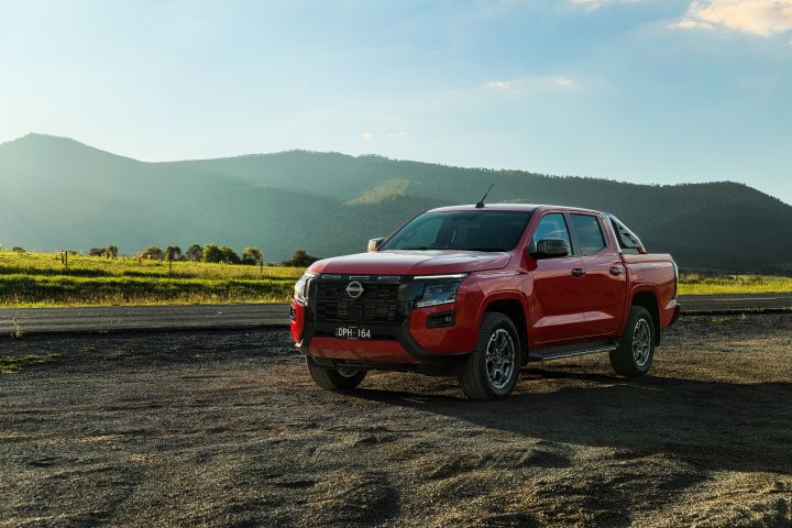 Front three-quarter view of a 2026 Nissan Navara ST‑X dual cab in Outback Red, parked on an open gravel surface with grassy fields and mountain ridgelines in the background.