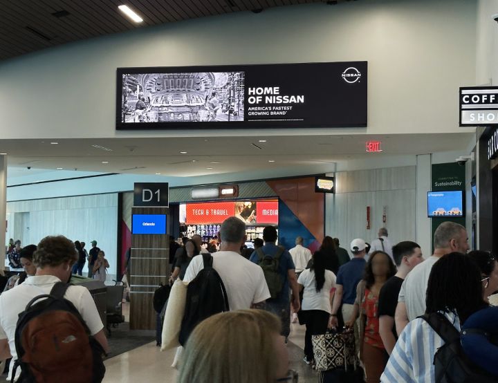 Travelers inside an airport, a "Home of Nissan" banner hangs on the wall.