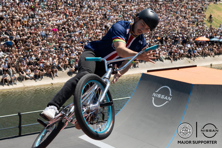 Male Bike Rider in the middle of the picture with crowd seating in the back, Nissan logo and FISE logo + “major supporter” mention on the botton right corner. / Homme bike rider au centre de la photo, foule assise en arrière-plan, logos Nissan et FISE en bas à droite de la photo.