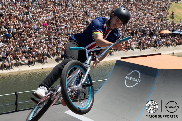 Male Bike Rider in the middle of the picture with crowd seating in the back, Nissan logo and FISE logo + “major supporter” mention on the botton right corner. / Homme bike rider au centre de la photo, foule assise en arrière-plan, logos Nissan et FISE en bas à droite de la photo.