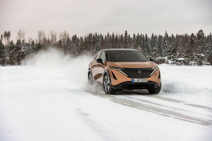 Elektroauto Nissan Ariya e‑4ORCE in Akatsuki Copper mit Dach und Außenspiegel in Pearl Black fährt auf Eissee mit schneebedeckten Bäumen im Hintergrund, 3/4 Front
