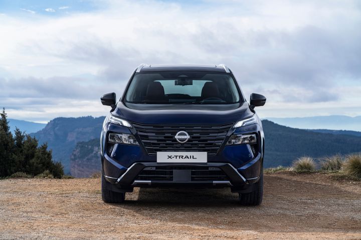 Neuer 2026 Nissan X‑Trail Tekna in der Außenfarbe Universal Blue, Frontansicht, geparkt auf unbefestigtem Untergrund mit bergiger Landschaft und bewölktem Himmel im Hintergrund. Das Fahrzeug verfügt über einen markanten Kühlergrill, schlanke Scheinwerfer und ein Nummernschild mit der Aufschrift „X‑TRAIL“.