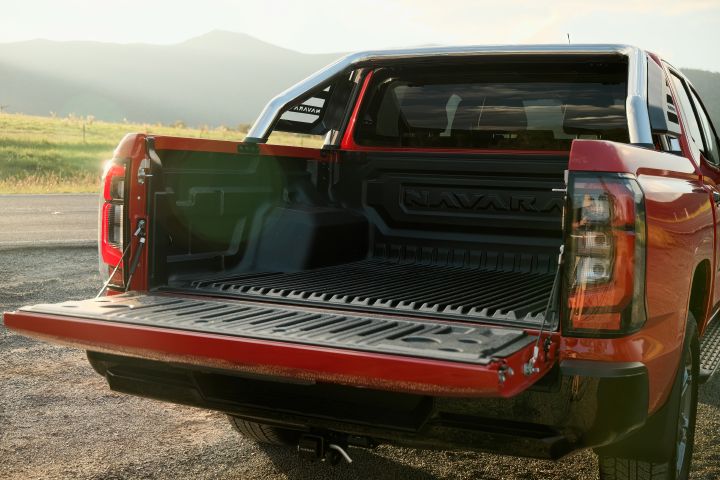 Rear three-quarter view of a 2026 Nissan Navara ST‑X dual cab in Outback Red with the tailgate lowered, showing the ribbed load floor, black bedliner, sports bar, and rear lamp cluster against an open rural landscape.