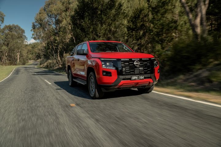 Front three‑quarter motion view of a 2026 Nissan Navara ST‑X dual cab in Outback Red, driving on a sealed road bordered by open grassy areas and trees.