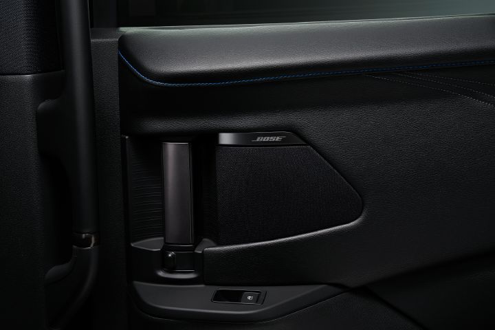 エルグランド AUTECHの後席スライドドア内張りをクローズアップで撮影した画像。画面中央にはブラックを基調としたドアトリムが広がり、上部には細かなシボ加工が施されたレザー調素材と、鮮やかな青色のステッチが横方向に伸びている。AUTECH専用インテリアを象徴するブルーステッチが、落ち着いたブラックの内装に上質なアクセントを加えている。
中央右側にはブラックのスピーカーグリルが配置され、その上部にはダーククロームの「BOSE」ロゴが確認できる。スピーカーの左側には縦向きのインサイドドアハンドル、さらにその左側には縦向きの大型グリップが備わり、後席乗員の乗降や車内移動をサポートする構造となっている。インサイドドアハンドルの下部にはドア開閉に関するスイッチが配置されている。
内装全体はブラックで統一され、レザー調素材、樹脂パネル、スピーカーグリルなど異なる質感が組み合わされている。
