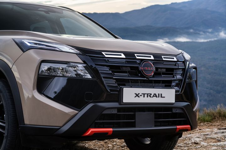 Vorderansicht des neuen 2026 Nissan X‑Trail, geparkt auf felsigem Gelände mit Bergen im Hintergrund. Zu den Merkmalen gehören ein markanter schwarzer Kühlergrill mit magmarotem Nissan-Emblem, schlanke LED-Scheinwerfer und magmarote Akzente an der unteren Stoßstange.