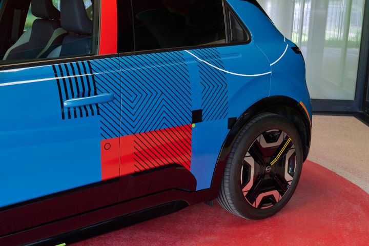 Nahaufnahme der Heckschürze des blauen Nissan Micra Signature Edition Gewinnerfahrzeugs mit schwarzen Zierstreifen, einem roten Akzentblock und einer großen Leichtmetallfelge vor einem roten Hintergrund